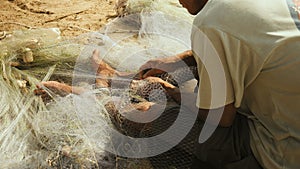 Close-up on a fisherman sitting down on the riverbank to mend a net by hand