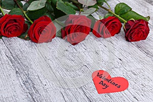 Red roses on a gray wooden background, with a heart with an inscription the day of St. Valentine, a free place for text.