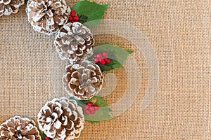 Close-up of flat lay Christmas wreath of pinecones, red berries, and holly leaves on rustic fabric background