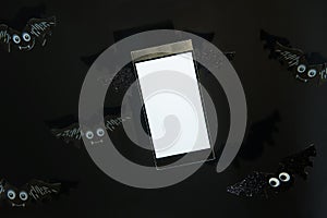 Close up flying smartphone with a blank white screen decorated with bat shape wings on the black background with bats