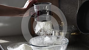 Close up footage of hand sifting white flour into glass bowl with a steel sieve.