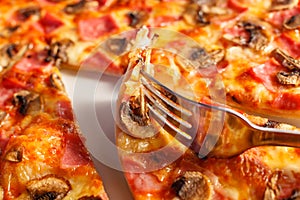 Close up fork takes a piece. Pizza with ham and mushrooms. Delicious hot food sliced and served on white platter. Menu