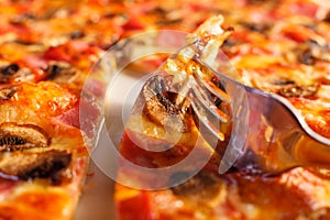 Close up fork takes a piece. Pizza with ham and mushrooms. Delicious hot food sliced and served on white platter. Menu