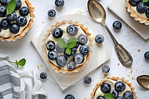 A close up of four blueberry tarts with mint leaves on top. The tarts are arranged on a white table with a silver spoon and a