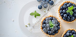 A close up of four blueberry tarts with mint leaves on top. The tarts are arranged on a white table with a silver spoon and a