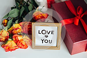 Close up frame with Love you message, gift box with red ribbon and a bouquet of roses flowers on the bedside