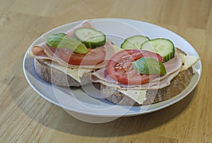 close up fresh home made rye bread sandwich with ham cheese sliced cucumbre tomatoes and basil leaves on white plate on wooden ta