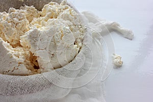 Close-up of freshly made white soft cottage cheese in bowl on white table background. Homemade ricotta in cheesecloth.