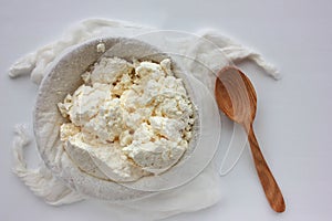 Close-up of freshly made white soft cottage cheese in bowl and wooden spoon on white table . Homemade ricotta in cheesecloth