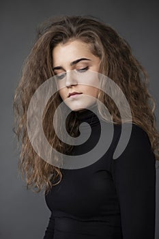Close up front portrait of a young woman with curly hair, wear black suitcase,  grey background, looking down.
