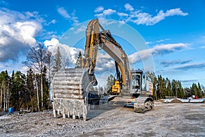 Close up front view at beautiful large yellow excavator with big scoop stands on new not yet ready road and new bridge. Sunny