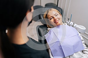 Close-up front view of happy young woman patient with dental problem talking with unrecognizable dental doctor during