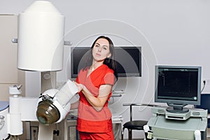Close up front view of high-skilled female doctor in red uniform, standing near the modern machine for non-invasive
