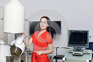 Close up front view of high-skilled female doctor in red uniform, standing near the modern machine for non-invasive