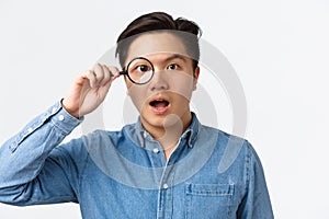 Close-up of funny asian guy looking through magnifying glass and look amazed, searching for something, found interesting