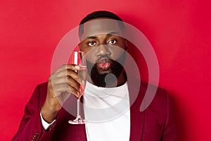 Close-up of funny Black man tasting champagne from glass, looking silly and pucker lips, celebrating new year, standing