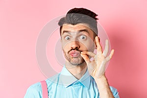Close up of funny caucasian guy touching his french moustache, pucker lips and looking silly, standing over pink