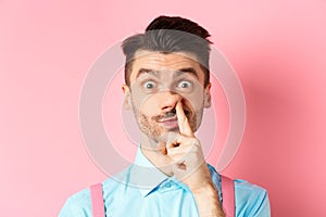 Close-up of funny caucasian man picking nose, staring silly at camera, standing on pink background