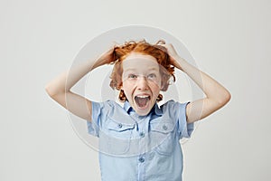 Close up of funny little boy with red hair and freckles pulling hair with hands,screaming with surprised expression