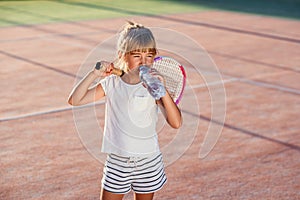 Close up funny little girl drinking water from bottle after hard tennis training at outdoor court at sunset. Sport