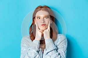 Close-up of funny redhead teen girl making faces, squinting and pocking cheeks, standing against blue background