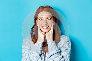 Close-up of funny redhead teen girl making faces, squinting and squeezing cheeks, standing against blue background