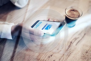 Close up of generic design smartphone with email sending icons screen holding in female hand. Cup espresso on the table