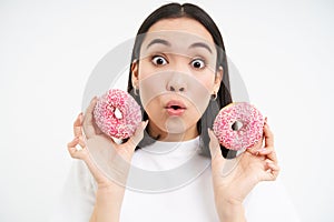 Close up of girl with amazed face, shows two glazed doughnuts, tempted to try new delicious donnut, white background photo