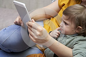 Close-up of a girl in her mother`s arms watching a tablet. Mom is working at home while taking care of her daughter. Concept of