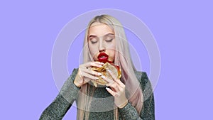 Close-up of a girl with red lipstick on her lips, eating a hamburger and smearing lipstick with her hand on a purple