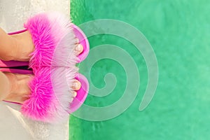 Close-up girl or woman feet in glamour fashion fluffy slippers standing on edge of pool with clear turquoise water. Legs wearing