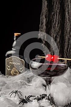 Close-up of glass of Halloween black widow cocktail with red cherries, spiders and dark bottle on spider web, black background