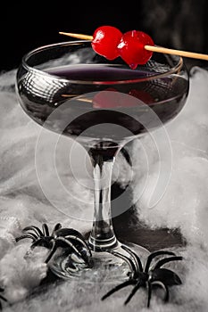 Close-up of glass of Halloween black widow cocktail with red cherries and spiders on spider web, black background,