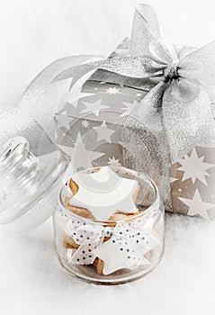 Close up of a glass jar full with christmas homemade star cookies over white fluffy background. Gift box in silver wrapping paper