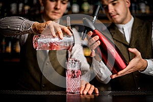 close-up on glass into which two bartenders simultaneously pour liquid from siphon and cup