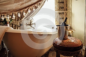 Close-up of a glass of wine and a bottle in an ice bucket on an elegant chair in the bathroom. A filled bubble bath next to the