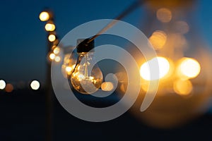 Close up of the glowing light bulbs at the beach cafe terrace. Sun is setting in the blurry abstract background.