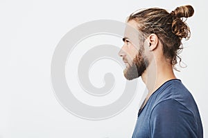 Close up of good-looking bearded hipster guy with hair in bun, in blue t-shirt standing in profile, looking aside