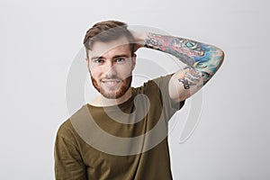 Close up of good-looking cool european guy with beard and arm tattoo smiling brightfully, holding hand behind head with