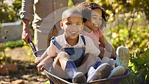 Close Up Of Grandchildren Pushed In Barrow By Grandfather Working In Vegetable Garden Or Allotment 