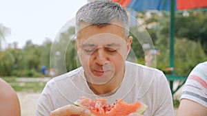 Close-up of a gray-haired man on a city beach eating watermelon with pleasure. Rest in the hot summer on the city beach