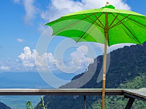 Close-up of green big umbrella and wooden table on beautiful blue sky and mountains background. Space for text. Concept of travel