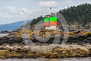 Close up of green and orange navigation marker on a rock in the Salish Sea, forested island and cloudy sky in background, San Juan