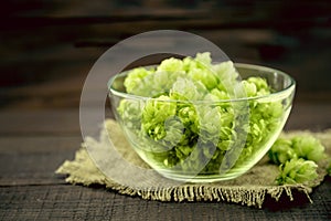 Close up of green ripe hop cones in a glass bowl over dark rustic wooden background. Beer production ingredient.