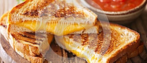 Close-up of grilled cheese sandwich with melting cheese, beside a bowl of tomato soup on a wooden board.