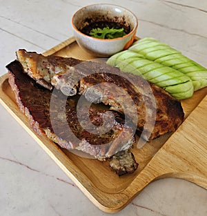 Close up Grilled pork ribs, sliced cucumber and spicy sauce on wooden tray.