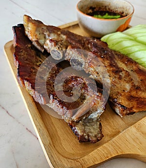 Close up Grilled pork ribs, sliced cucumber and spicy sauce on wooden tray. Barbecue food with fresh vegetables on white marble.
