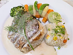 Close-up grilled sea bass steak served with white wine sauce, fried rice with butter, and cooked vegetables on white plate.