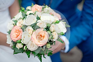 Close-up groom in a suit and the bride in a white dress are standing and holding a bouquet of peach roses, eustomas and