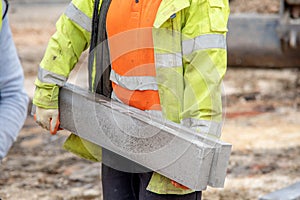 Close up of groundworker in orange and yellow hi-viz  carrying heavy concrete kerbs on construction site during new road